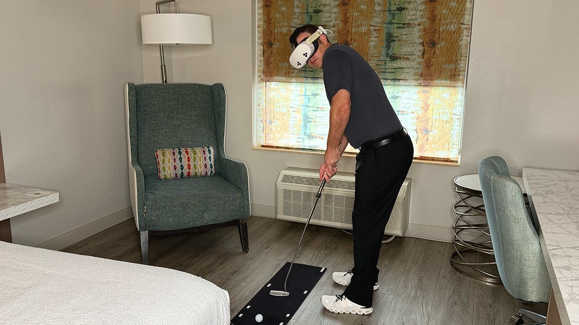 A golfer in a Meta Quest headset putting off a dark mat in a hotel room, beside the bed, with a real putter and ball.