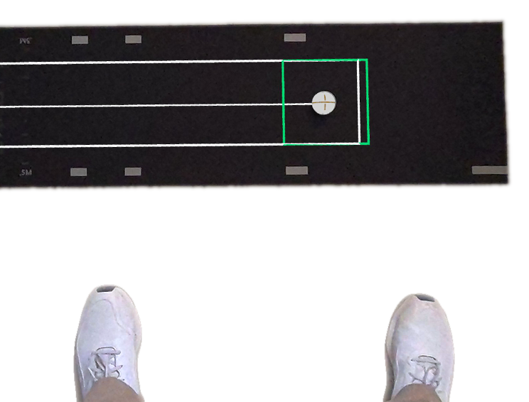 Looking down from address: the dark mat ahead with the alignment line along it, the virtual ball in a green zone inside the white box, and the golfer's shoes in frame.